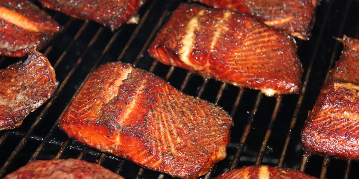 Perfectly smoked salmon fillets with mahogany-red crust and smoke rings cooking on dark metal smoker grates, demonstrating the final result of proper fish brining technique