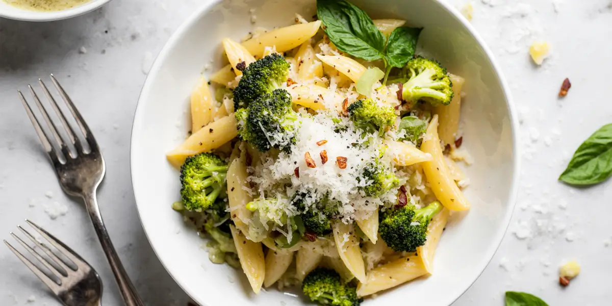 Light broccoli pasta recipe in white bowl with fresh green broccoli florets, penne pasta, olive oil, Parmesan cheese and basil garnish on marble countertop