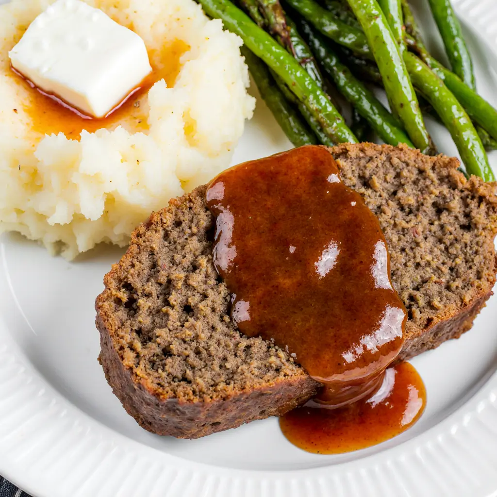 Classic meatloaf slice served with mashed potatoes and green beans on a white plate, topped with a shiny ketchup glaze.